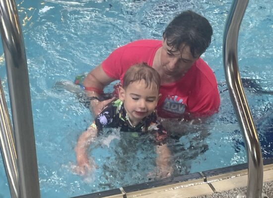 Swimming instructor in pink top holding three year old boy by pool steps